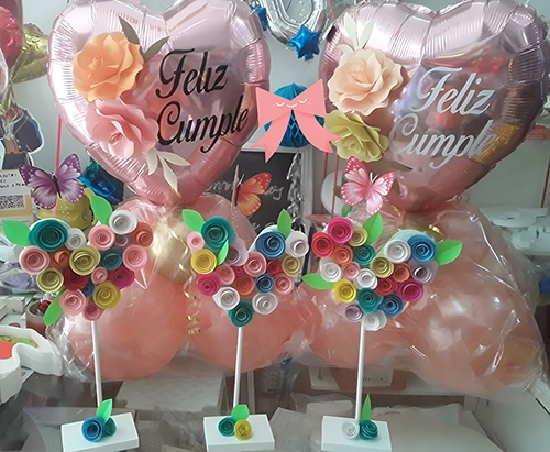 Bouquet festejo de cumpleaños. Se ven dos globos corazón rosa metalizado cada uno con la palabra Feliz Cumple estampada en blanco y en negro. Delante, tres corazones más pequeños, armados con flores de cartulina y varilla a modo de tallo en base de madera.